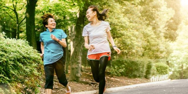 Two women in their 50s jog down a wooded trail laughing with each other.