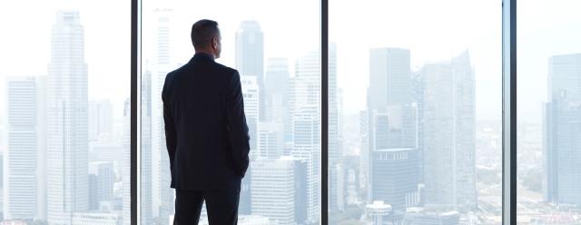 standing man in suit looking out tall window of high rise